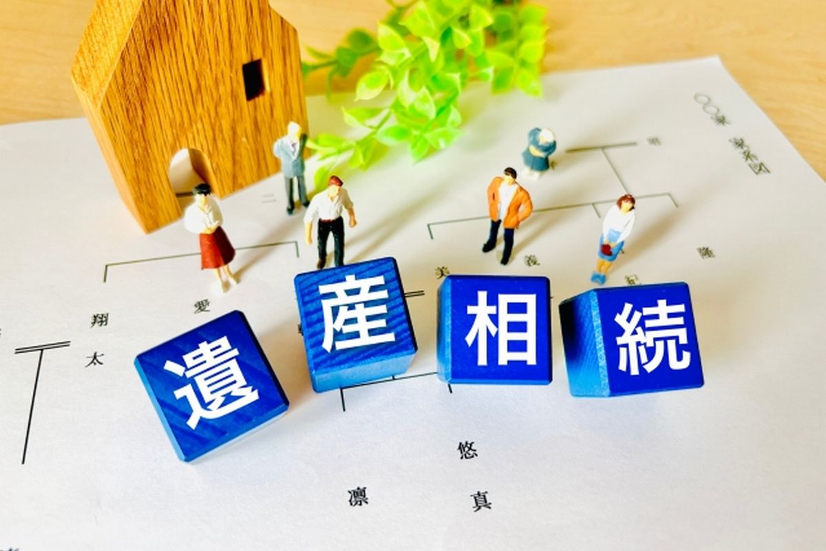 机上の家系図の上に、人々のフィギュアと家の模型、そして「遺産相続」と書かれた積み木が置かれているイメージ。兄弟間の相続や遺産分割を象徴する写真。